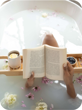 Mulher em uma banheira com pétalas de rosa, com apenas mãos e pernas aparentes, segurando um livro com uma bandeija com uvas e chá